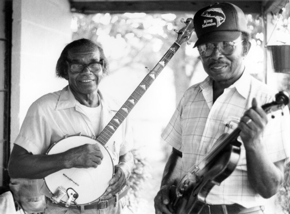 Two Black fiddlers, one with a banjo and the other with a violin, smiling at the camera. King Salmon cap.