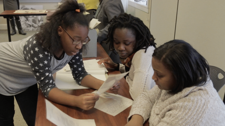 Three Black women reviewing documents at a table, focusing on Black girlhood.