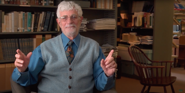 Man with a beard in a library setting, gesturing while seated.