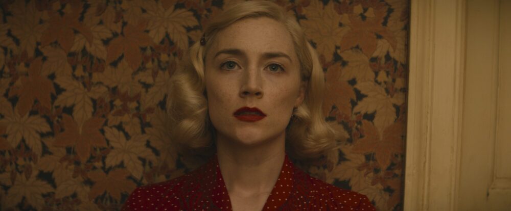 Saoirse Ronan in "Blitz," a film set against patterned wallpaper, wearing a red polka dot dress and red lipstick.