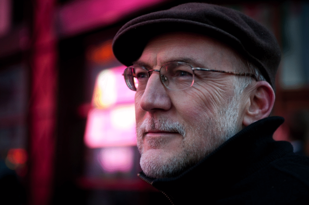 Portrait of Bob Shaw wearing a cap and glasses, with a blurred, colorful background.