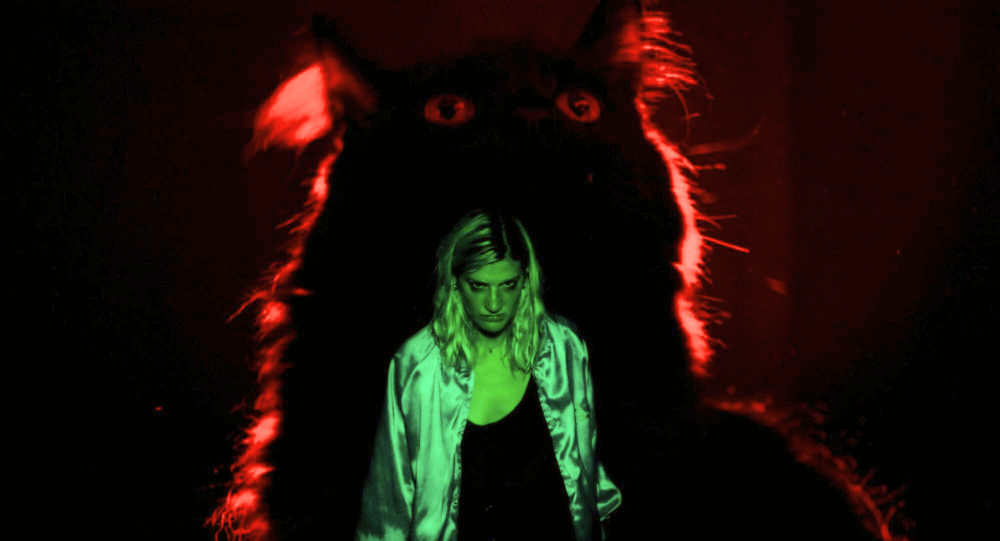 Woman with green lighting with a large cat with red lighting behind her.