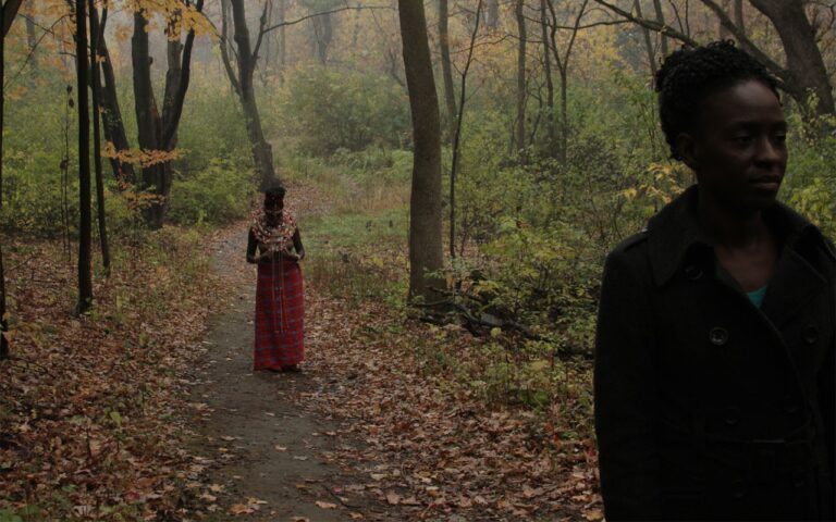 Woman in coat with a figure in traditional African dress in a wooded area.