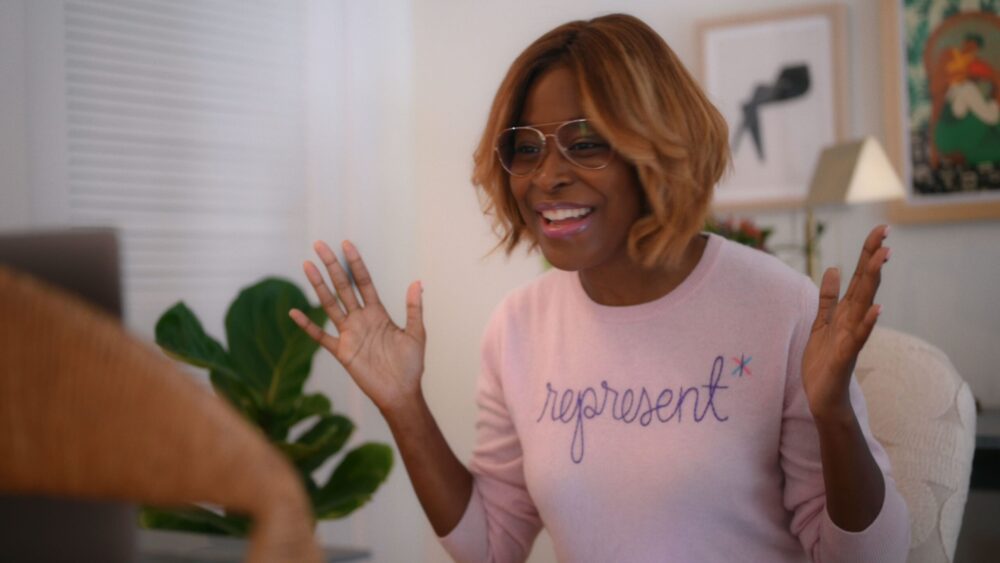 Enthusiastic woman wearing a "represent" sweater and glasses, gesturing with hands.