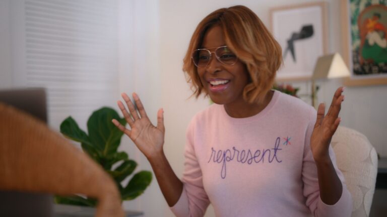 Enthusiastic woman wearing a "represent" sweater and glasses, gesturing with hands.