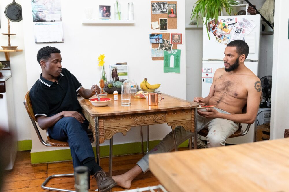 Two men eating at a table in an apartment. "Before you play.