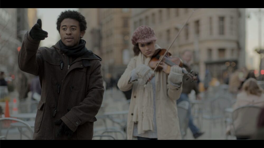 Man gestures near a violinist playing on a city street.