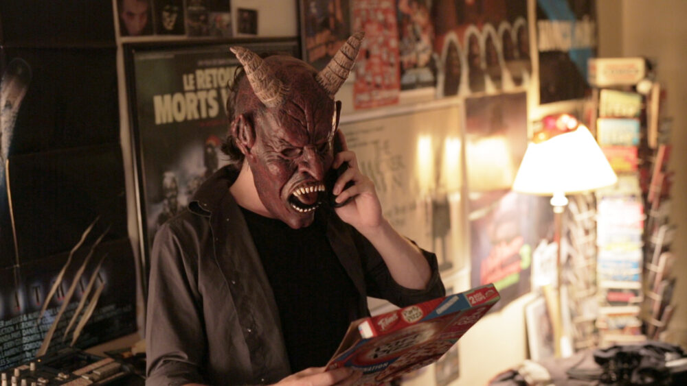 Man in a devil mask on the phone, holding a pizza box in a room with horror movie posters.