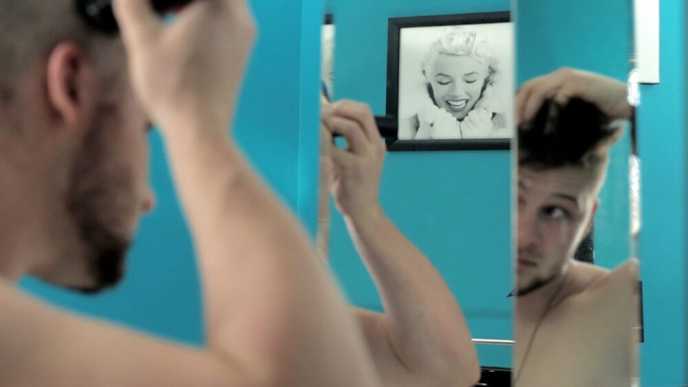 Man giving himself a haircut in front of a mirror, with a Marilyn Monroe picture in the background.