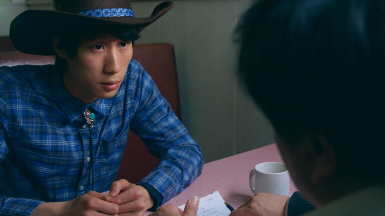 Young man in cowboy hat and blue plaid shirt at a diner table.