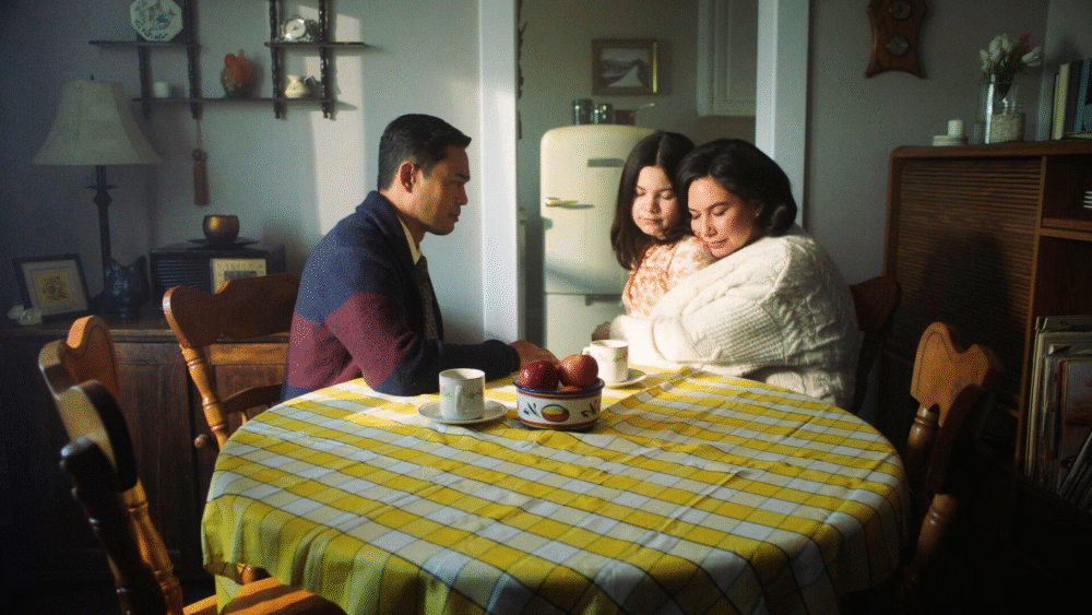 Family gathered around a table with a yellow tablecloth, embracing in a scene from "Café Daughter.
