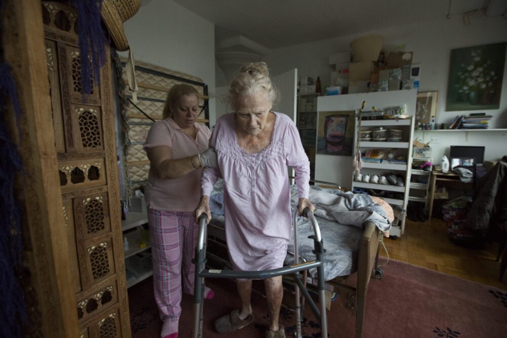 Caregiver assisting elderly woman with walker in bedroom.