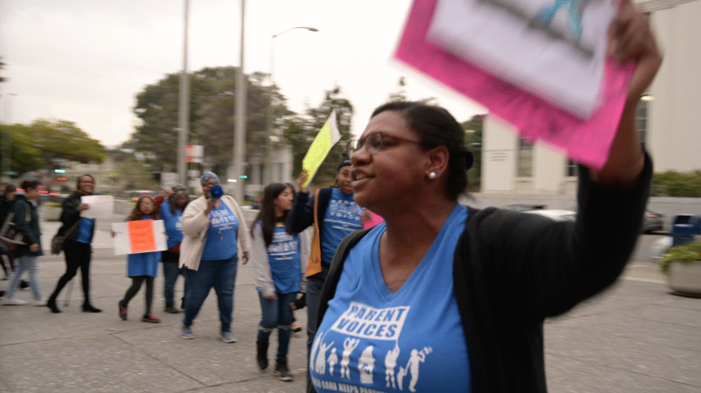 Parents protesting, wearing "Parent Voices" shirts, holding signs.