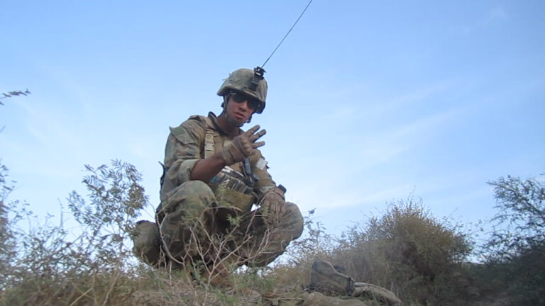 Soldier in combat gear squats in a field.