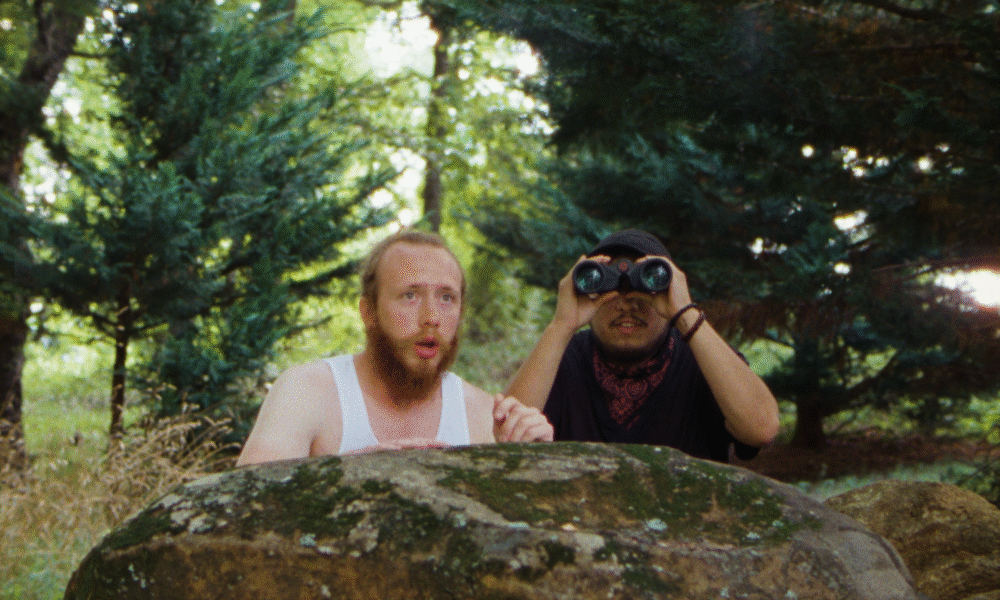 Two men behind a rock, one using binoculars in the woods.