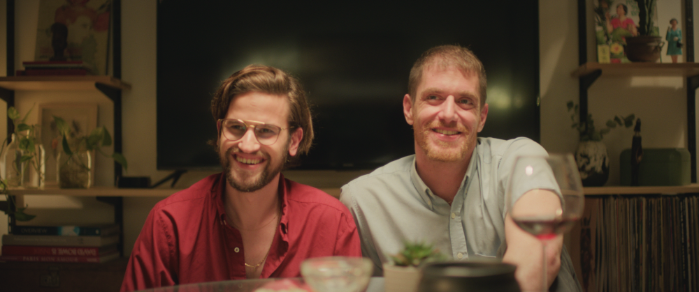 Two men smile at the camera in a cozy living room setting.
