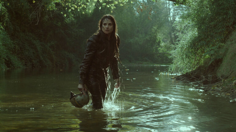 Woman in wet clothes wading through a river, holding a helmet.