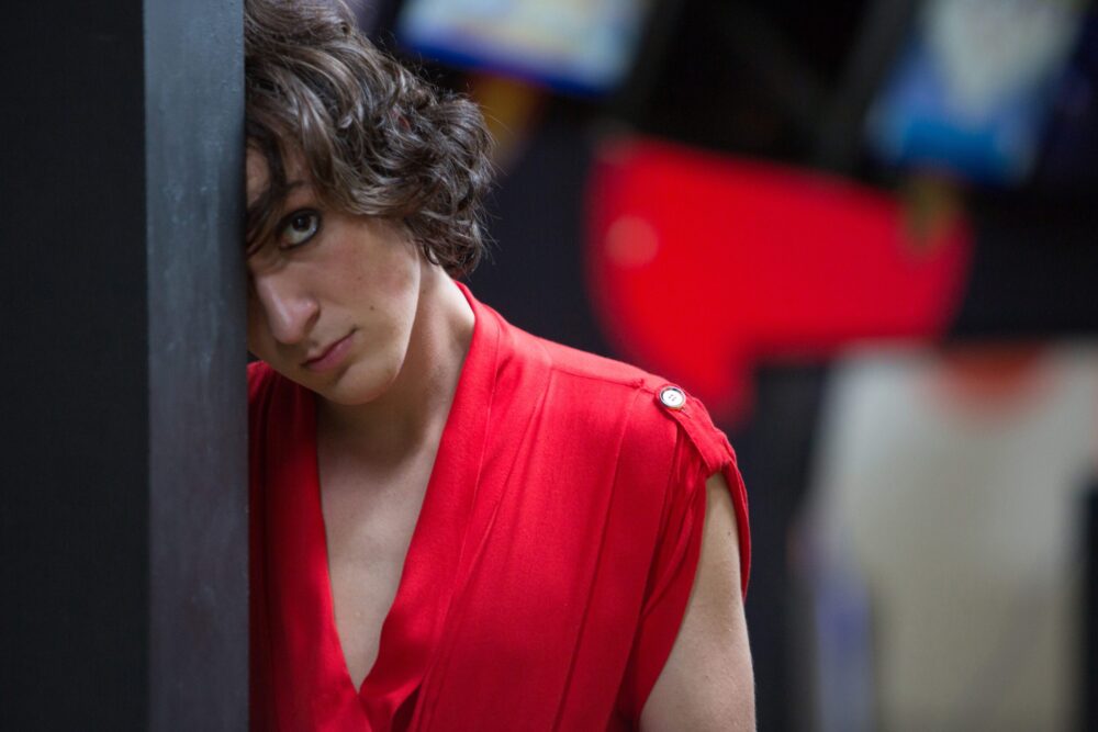 Intense young person in red shirt leaning against a wall.