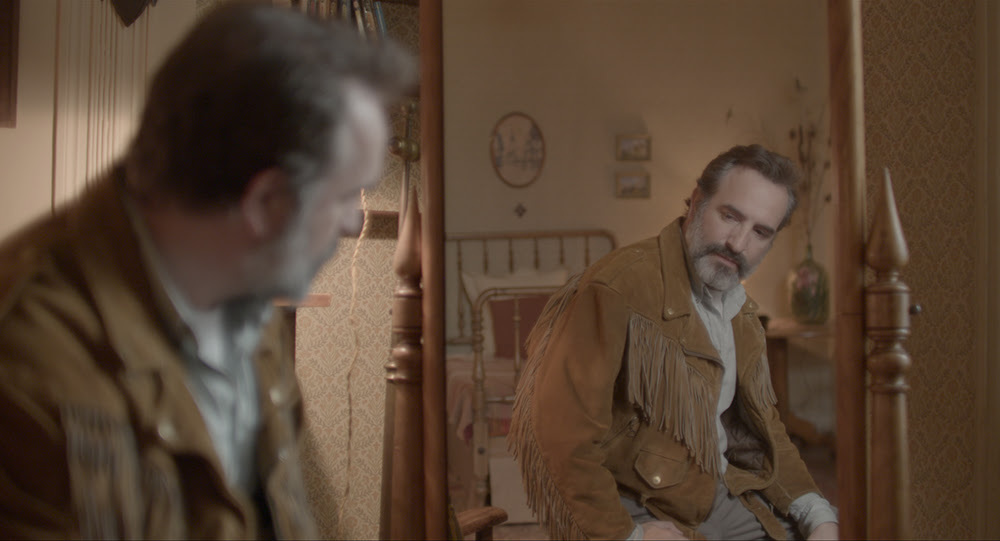 Man in a fringed jacket looking in a mirror.