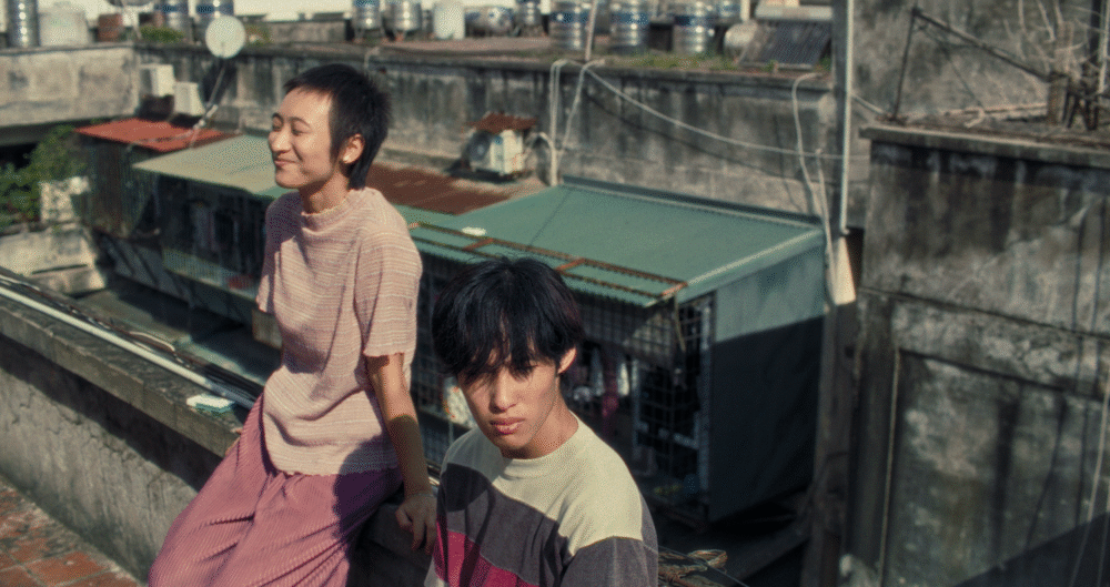 Two young people on a rooftop, one sitting and smiling, the other looking at the camera.