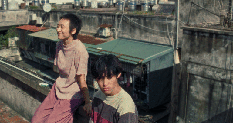 Two young people on a rooftop, one sitting and smiling, the other looking at the camera.