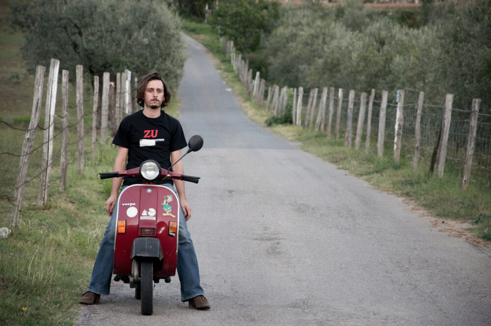 Man on a red Vespa scooter on a rural road with a fence.