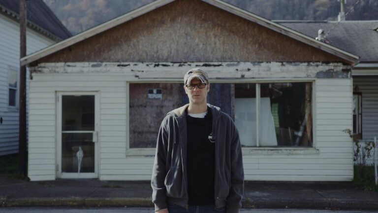 Man stands in front of weathered storefront, "Devil Put the Coal in the Ground