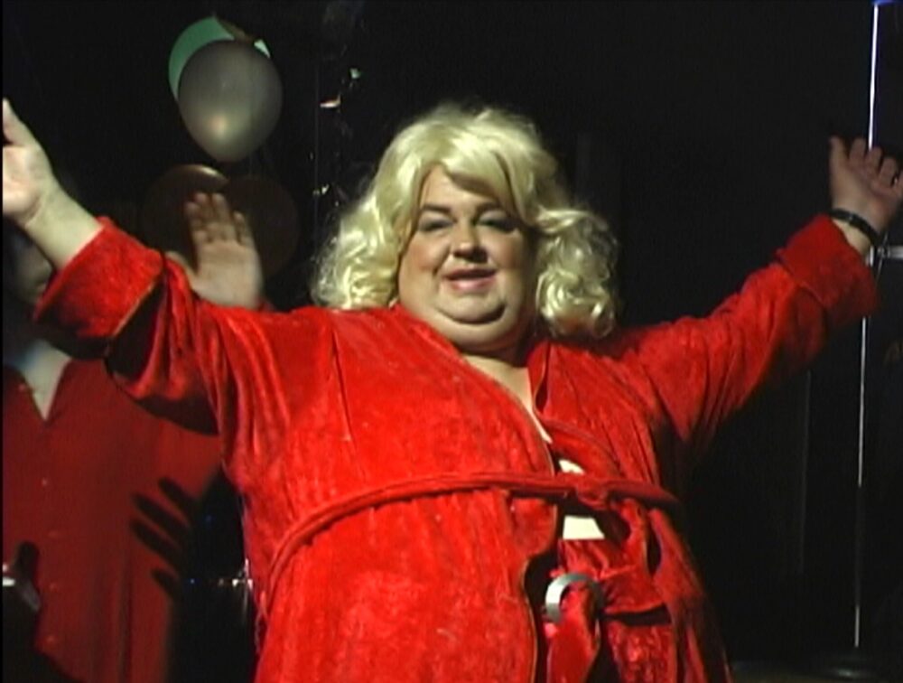 Dirtwoman documentary image: A person in a red robe and blonde wig smiles with arms raised.