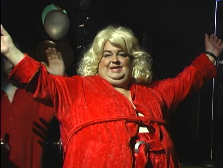 Dirtwoman documentary image: A person in a red robe and blonde wig smiles with arms raised.