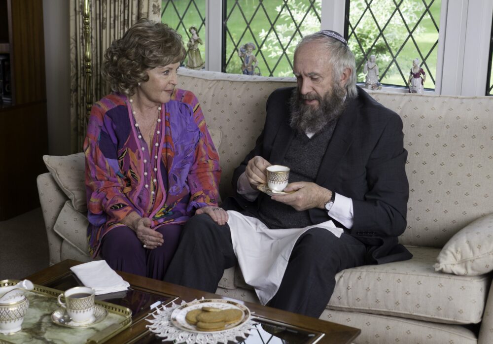 Elderly couple on sofa, man with kippah holds tea.