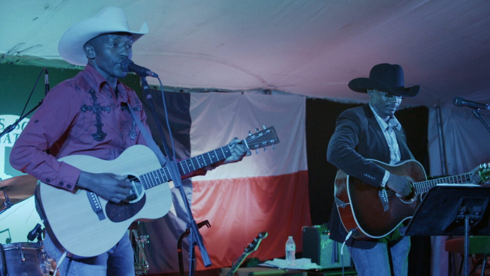 Dusty & Stones perform live: Two Black men in cowboy hats playing guitars on stage with a Texas flag backdrop.