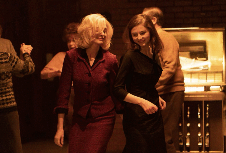 Two women dance near a jukebox, one in a red suit, the other in black. Scene from "Eileen".