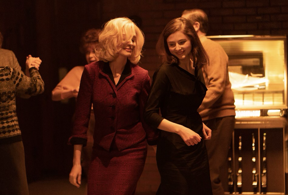 Two women dance near a jukebox, one in a red suit, the other in black. Scene from "Eileen".
