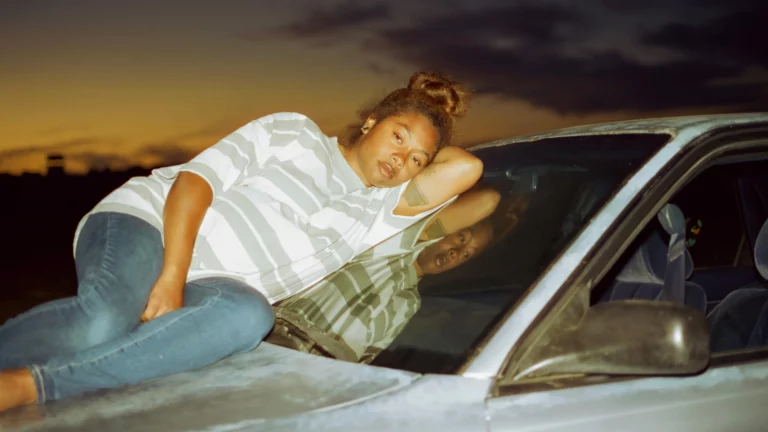 Woman lying on car hood at sunset, reflection in windshield. Earth Mama aesthetic.
