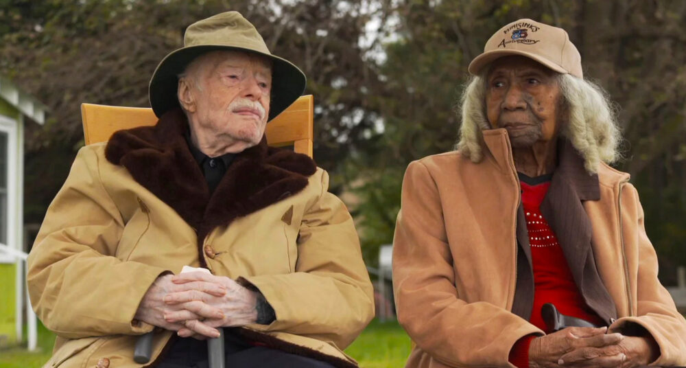 Two elderly people sit outside, one wearing a "Ministry 25 Anniversary" cap.