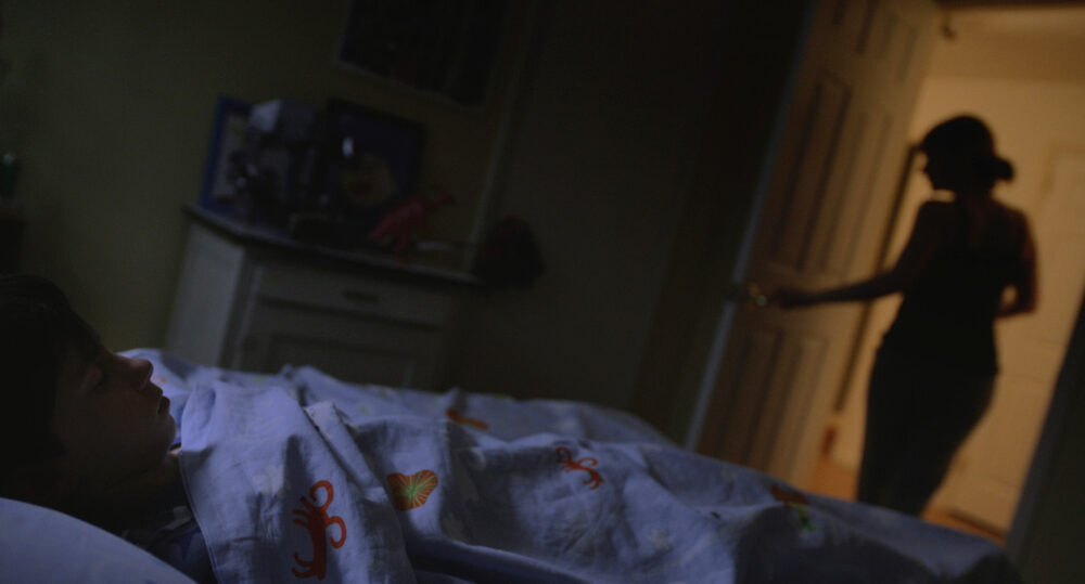 Child asleep in bed, with a silhouette of a woman in the doorway, hinting at an empty bed.