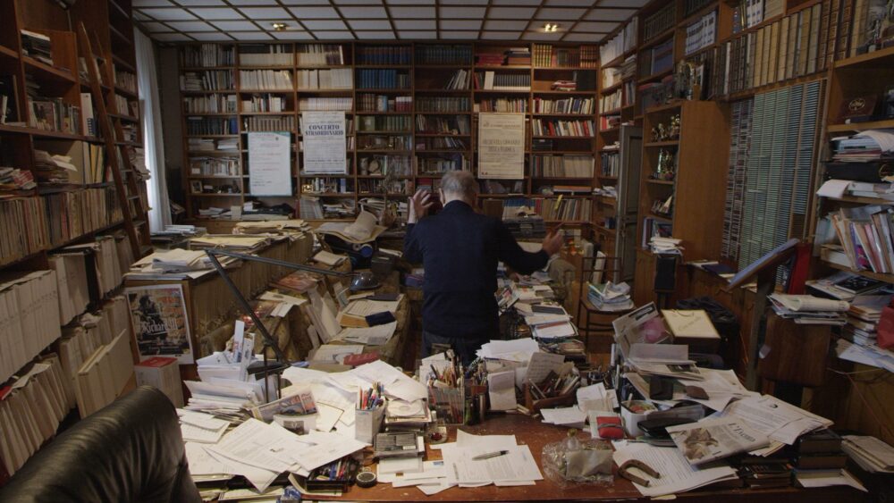 Ennio Morricone conducting in his cluttered home studio surrounded by books and papers.