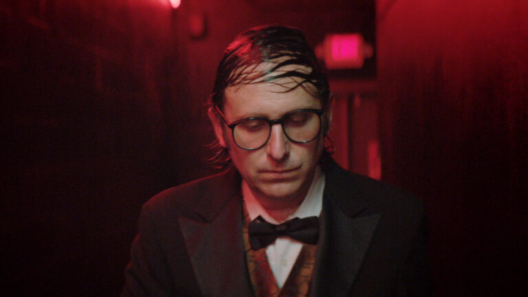 Man in tuxedo and glasses under red lighting, looking down.
