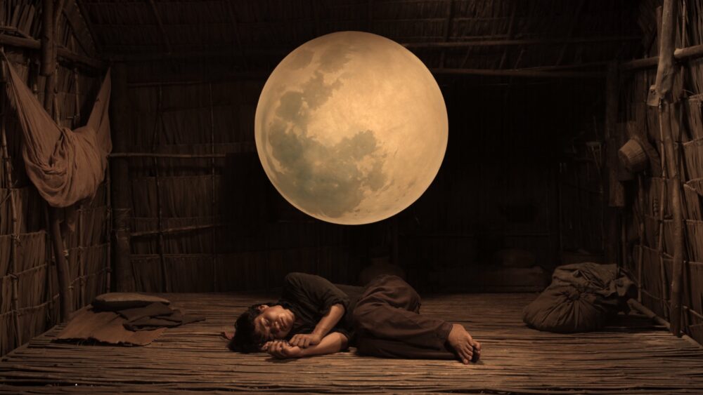 Person sleeping under a large moon in a rustic hut.