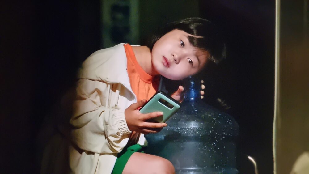 Young child with phone leaning on water jug, wearing jacket and shorts.
