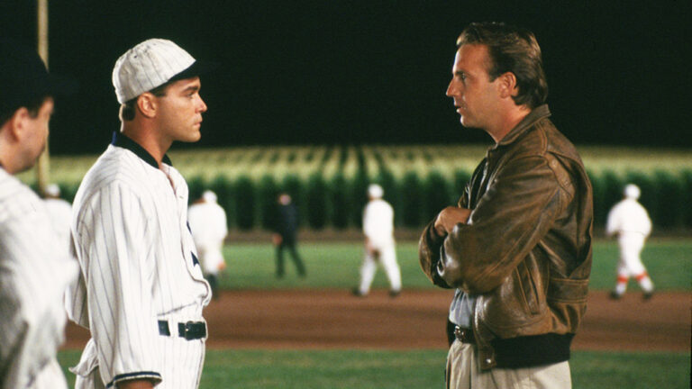Kevin Costner in Field of Dreams talking to a baseball player on a baseball field at night.