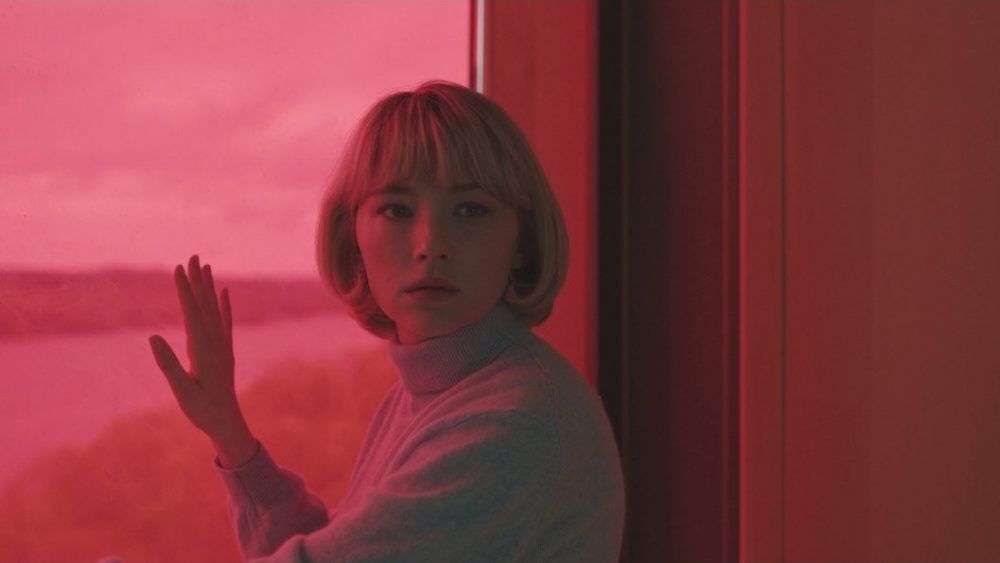 Haley Bennett in "Swallow" movie, looking out a pink-tinted window with a worried expression.