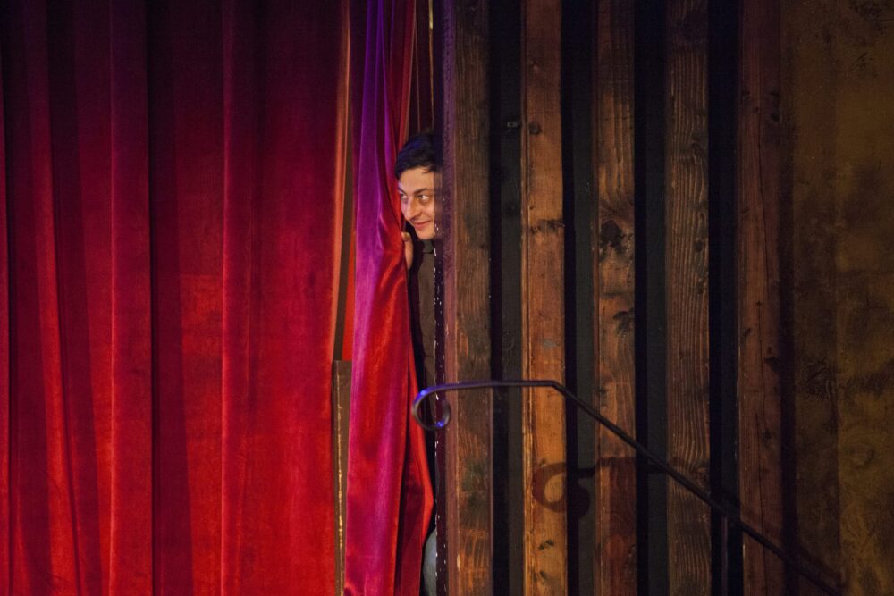 Comedian peeking from behind a red curtain and wood paneling.