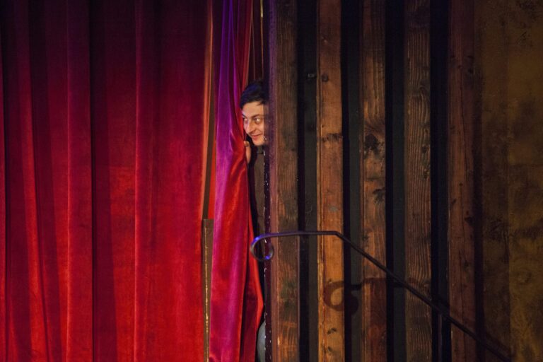 Comedian peeking from behind a red curtain and wood paneling.