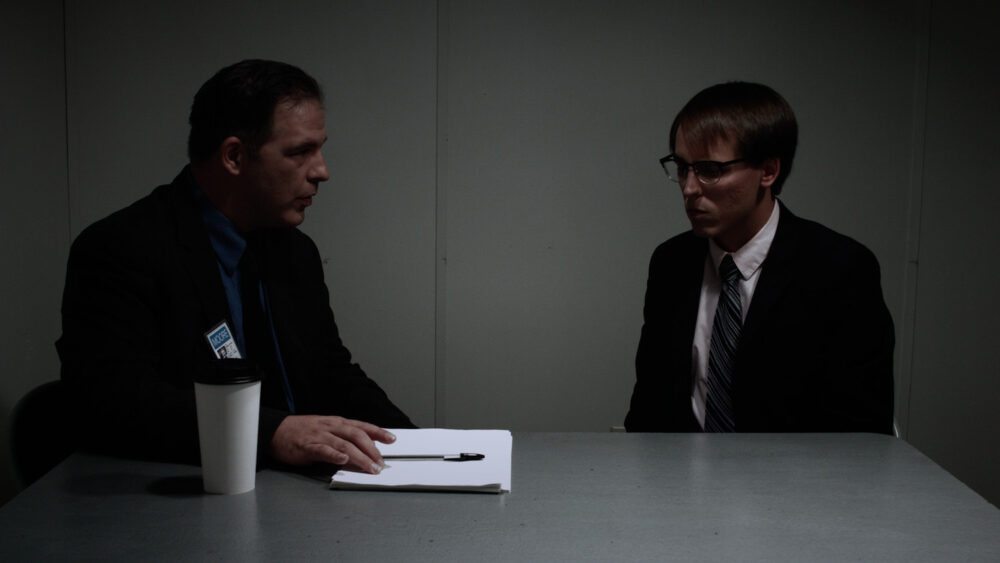 Interrogation scene: Detective questions a man in a suit across a table.