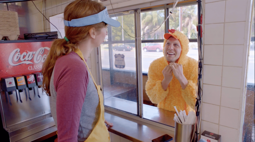 Woman at food place window serving a customer in a chicken costume.