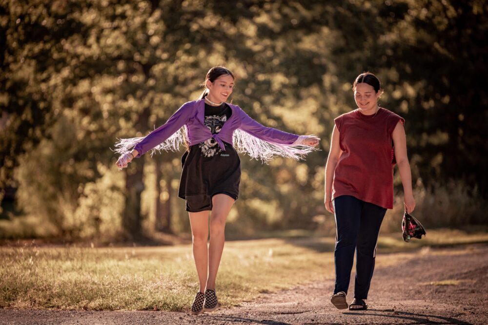 Two girls walking on a path in "Fancy Dance" attire. One is wearing a purple fringed jacket.