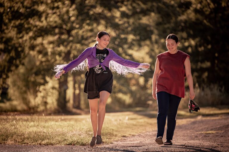 Two girls walking on a path in "Fancy Dance" attire. One is wearing a purple fringed jacket.