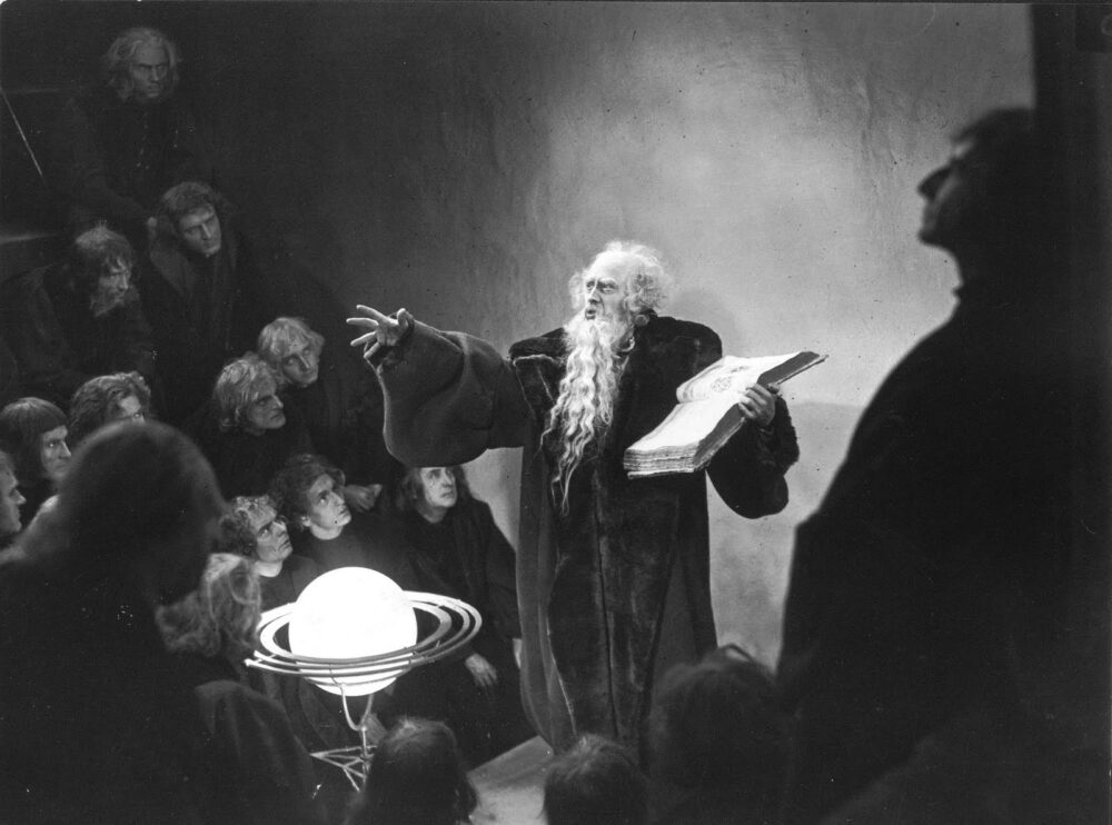 Scene from "Faust": A robed figure gestures while holding a book, surrounded by onlookers and a model of a planet.