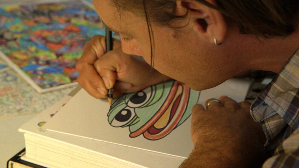 Artist drawing Pepe the Frog, a meme symbolizing "Feels Good Man," on paper with colored pencils.